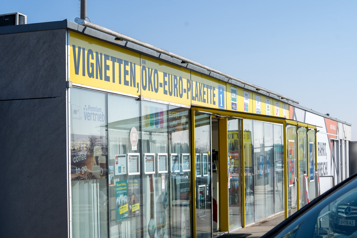 Austrian Premium-Vertrieb sales kiosk near Bratislava border with bold yellow 'VIGNETTEN, ÖKO-EURO-PLAKETTE' signage, selling vignettes for multiple countries plus German Umweltplakette, ASFINAG branding visible, Austrian plate WD-119 AZ in foreground, February 2026