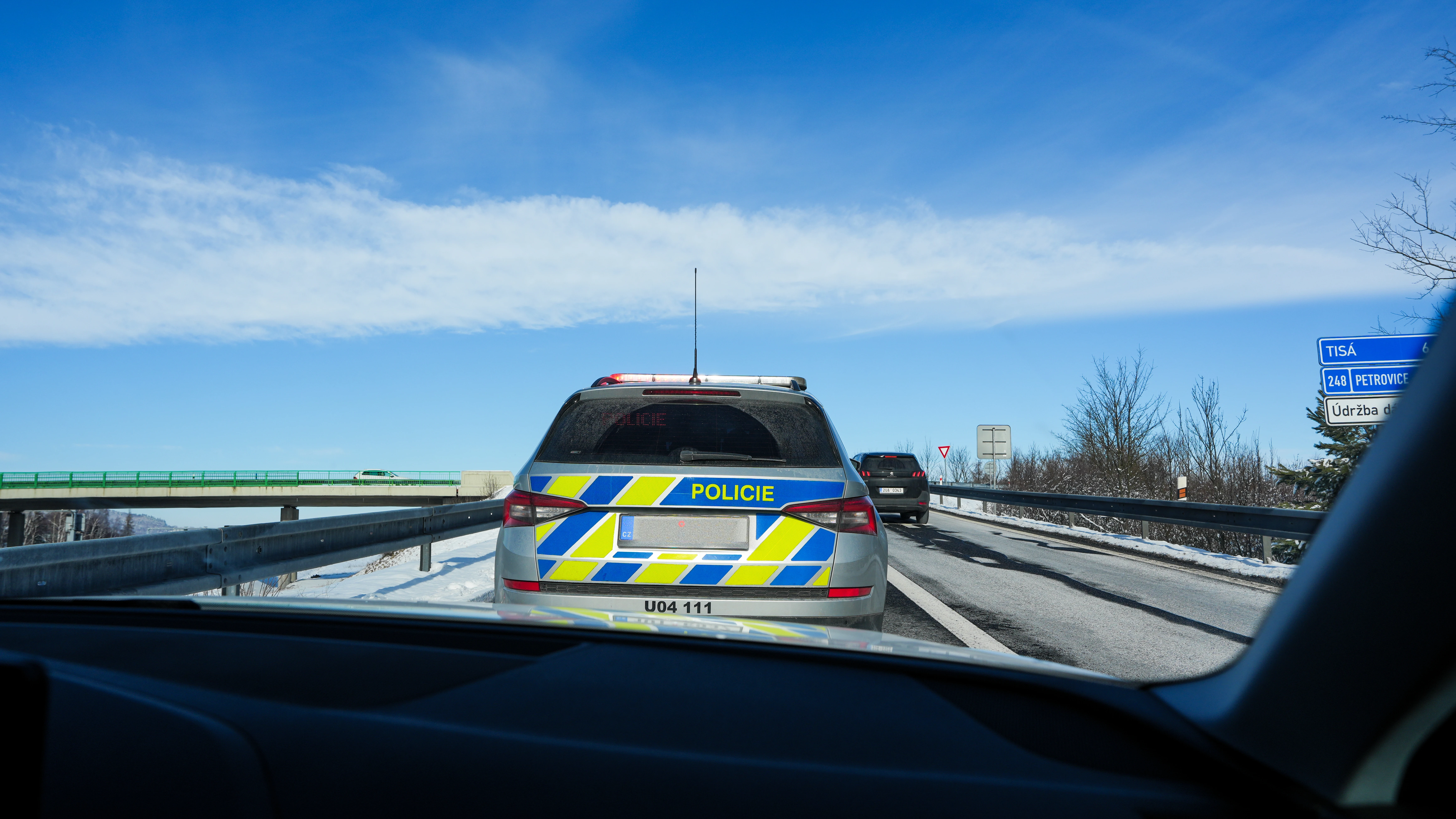  Czech Policie Škoda patrol car on D8 motorway in winter with snow on roadside, Tisá and Petrovice direction signs visible, photographed from following vehicle, February 2026