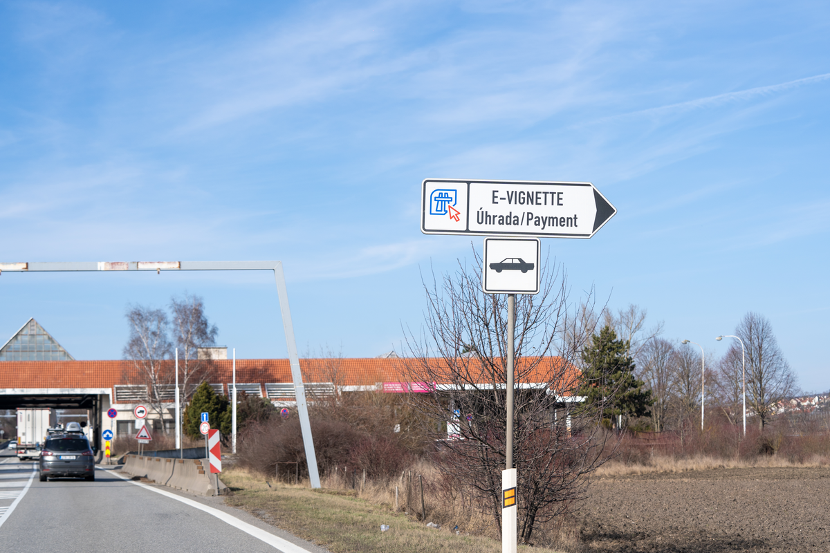 Bilingual Czech-English directional sign reading E-VIGNETTE Úhrada/Payment with right-pointing arrow and car symbol, directing drivers to an e-vignette sales kiosk at a Czech border crossing approach, with former border post buildings and traffic visible behind, February 2026