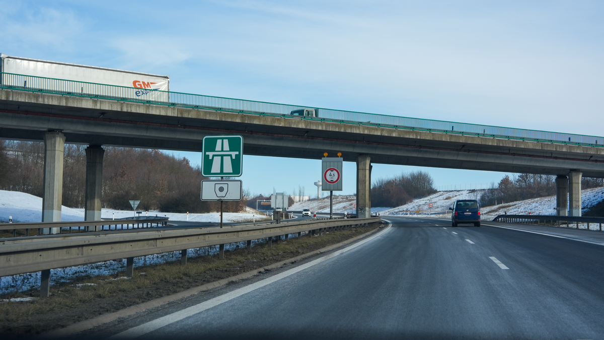 Czech motorway entry sign with green motorway symbol and vignette icon below it marking where the e-vignette obligation begins, overpass bridge with HGV truck passing overhead, 4.5m height restriction sign, snow-dusted verges and clear blue winter sky on D8 motorway, February 2026