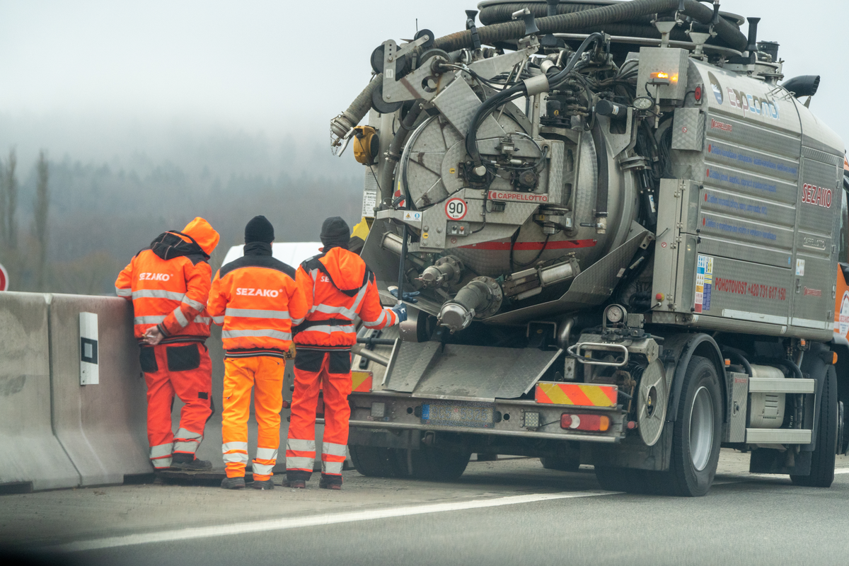  Three SEZAKO road maintenance workers in high-visibility orange overalls working alongside a Cappellotto vacuum cleaning truck with Czech 9C5 5325 plates on D5 motorway in foggy winter weather, performing drainage maintenance work, vehicle exempt from Czech e-vignette requirement, February 2026