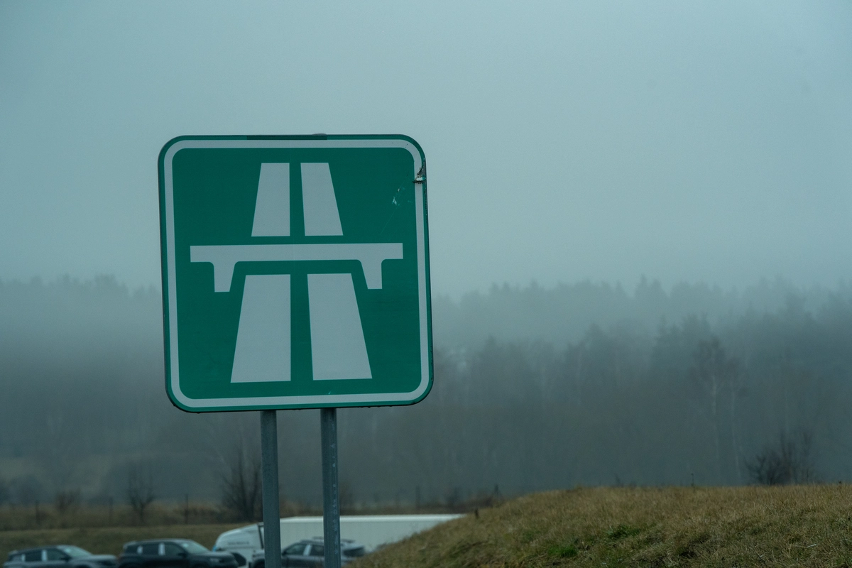 Czech motorway entry sign with vignette exemption symbol (e-vignette icon crossed out) at start of a vignette-free motorway section, winding road through snow-covered pine forest with red-and-white chevron markers on the bend, clear blue winter sky, February 2026