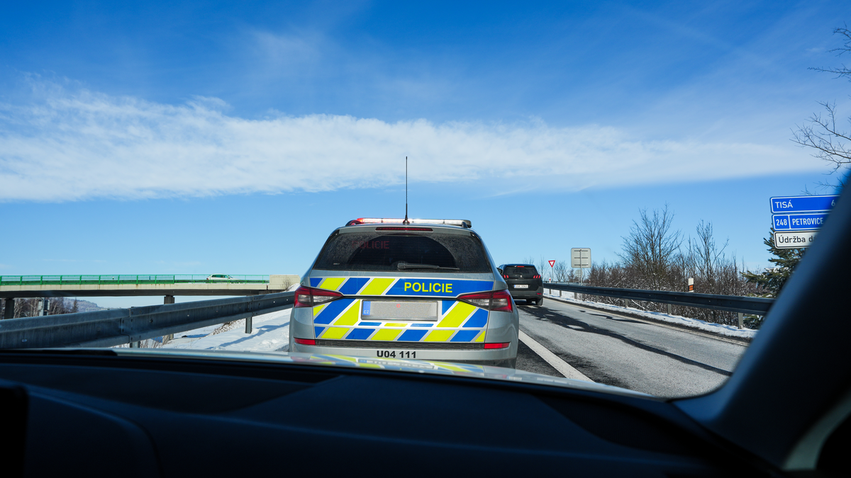 Czech Policie Škoda patrol car on D8 motorway in winter with snow on roadside, Tisá and Petrovice direction signs visible, photographed from following vehicle, February 2026