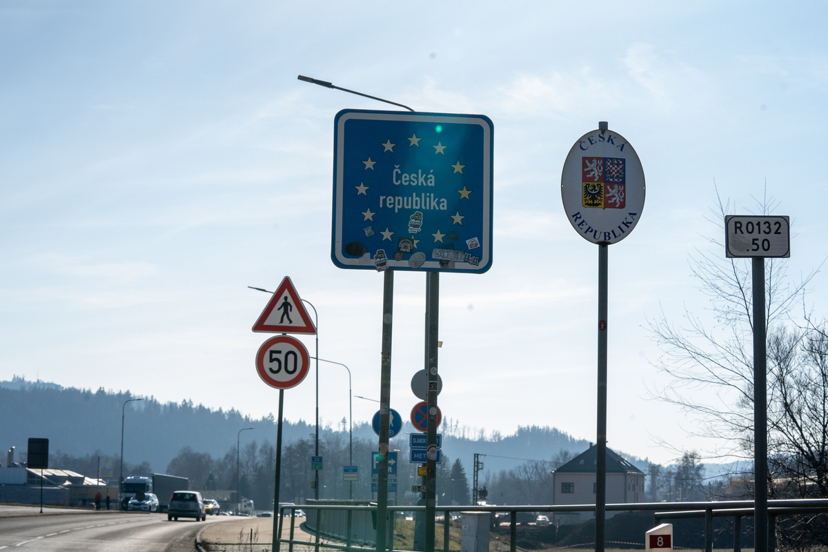 Blue EU-style Česká republika border entry sign covered in travel stickers next to white oval Czech coat of arms sign at the Náchod–Kudowa-Zdrój crossing on the E67/I/33 between Czech Republic and Poland, with 50 km/h speed limit and direction signs to Náchod visible, February 2026