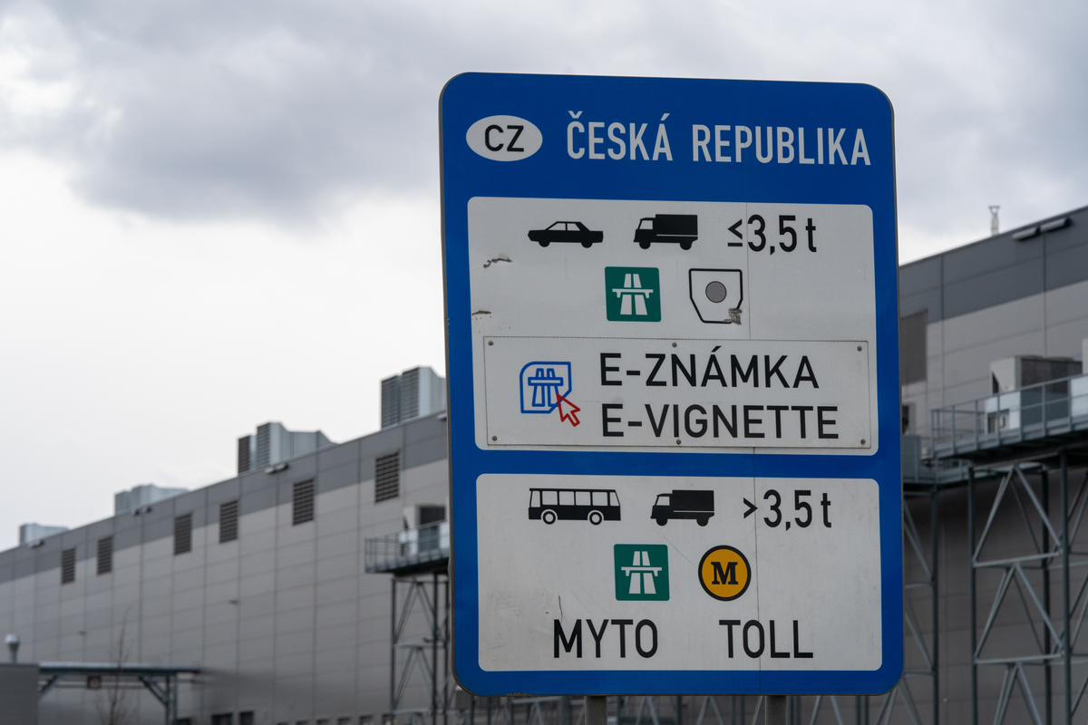 Czech Republic border information sign at the Czech–Slovak border showing e-vignette obligation for vehicles ≤3.5t with bilingual Slovak/English 'E-ZNÁMKA / E-VIGNETTE' wording and MYTO TOLL for vehicles >3.5t, industrial warehouse buildings in the background, March 2026
