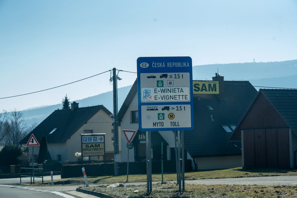 Czech Republic border information sign with bilingual Polish/English E-WINIETA / E-VIGNETTE wording for vehicles ≤3.5t and MYTO/TOLL for vehicles >3.5t, with Polish SAM Spożywczy and Czech Potraviny grocery store signage and mountain village in the background near the Czech–Polish border, February 2026