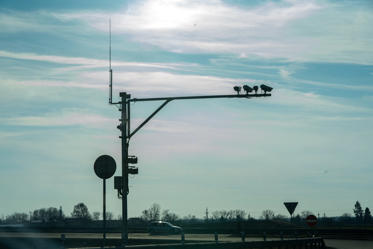 Cantilever-mounted Czech vignette enforcement gantry with four ANPR cameras and antenna silhouetted against late afternoon sky at the Czech–Slovak border, single car passing below with no-entry and give-way signs visible, February 2026