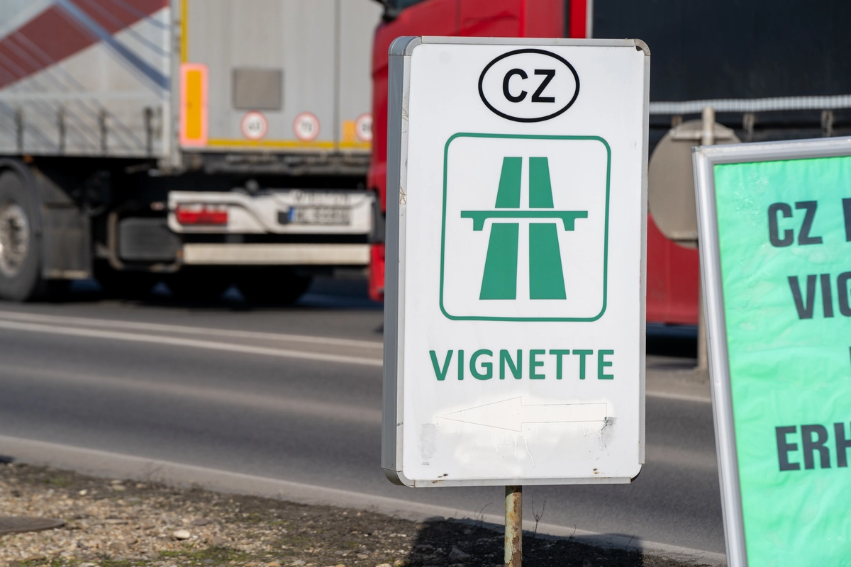 Czech vignette roadside sales sign with CZ oval, green motorway pictogram, 'VIGNETTE' wording and white directional arrow at the Dolní Dvořiště border crossing, German HGV passing in the background, February 2026
