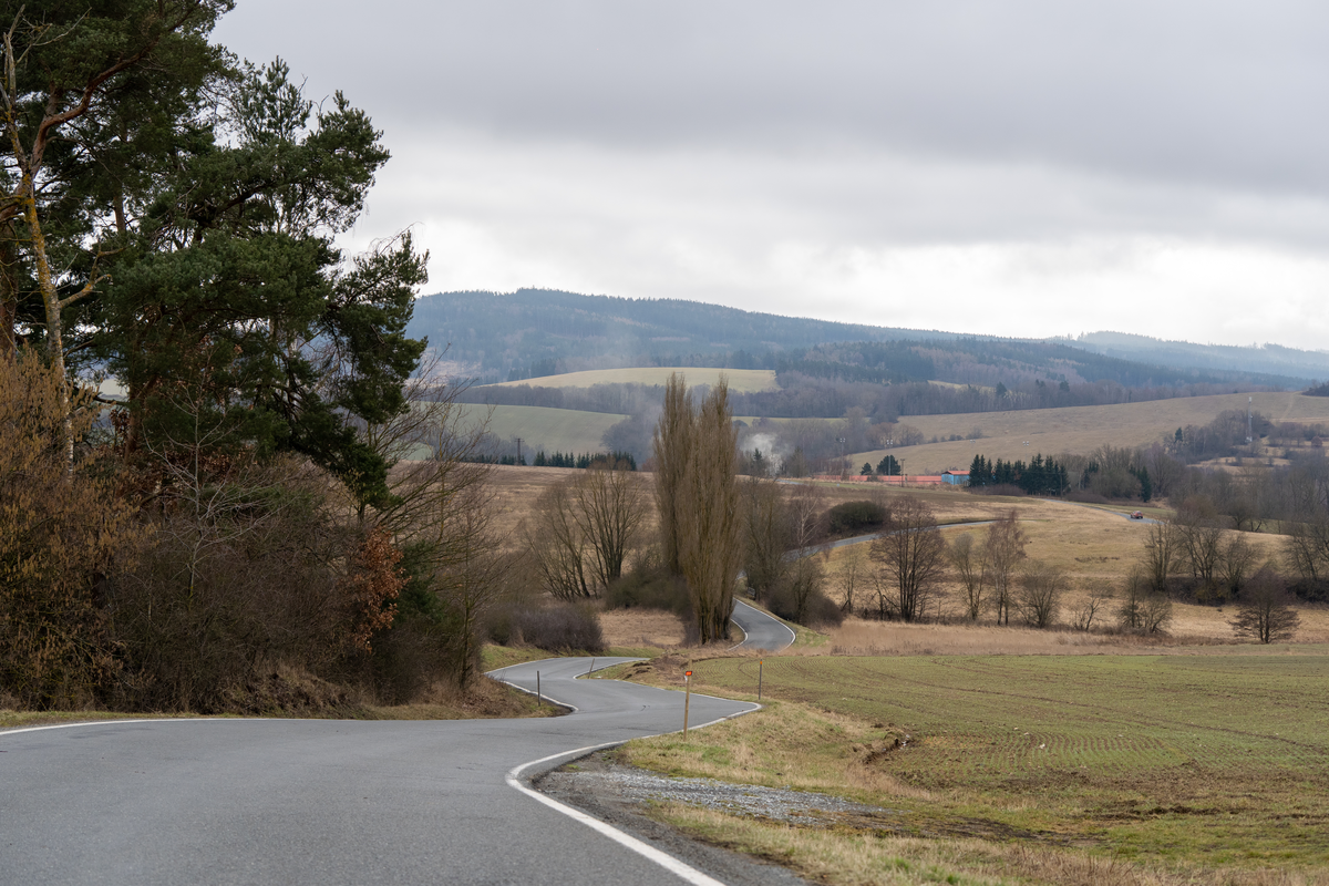 Quiet winding Czech country road meandering through rolling hills and fields with bare poplar trees, distant forested mountain range and overcast late winter sky in west Bohemia near the Plzeň region, vignette-free class III road, February 2026
