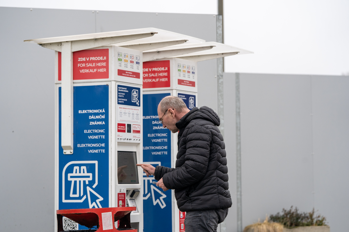 Kierowca kupujący czeską e-winietę w samoobsługowym kiosku z e-winietą edalnice.cz w miejscu obsługi podróżnych (MOP) przy czeskiej autostradzie niedaleko przejścia granicznego w Rozvadovie, z trójjęzycznym oznakowaniem czesko-angielsko-niemieckim pokazującym warianty winiety: roczny, 30-dniowy, 10-dniowy i 1-dniowy, luty 2026