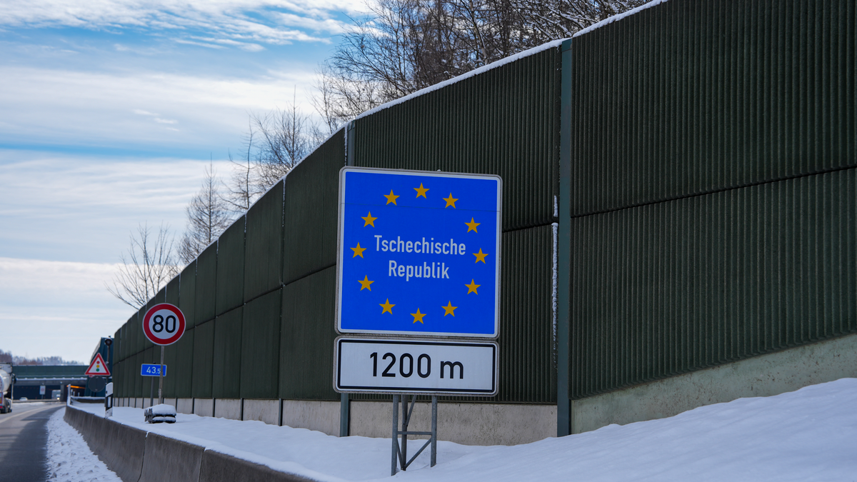 German EU-style Tschechische Republik advance warning sign indicating the Czech Republic border 1200 metres ahead on the A17 autobahn from Dresden, with 80 km/h speed limit and snow-covered noise barriers, approaching the Petrovice–Breitenau crossing in winter, February 2026