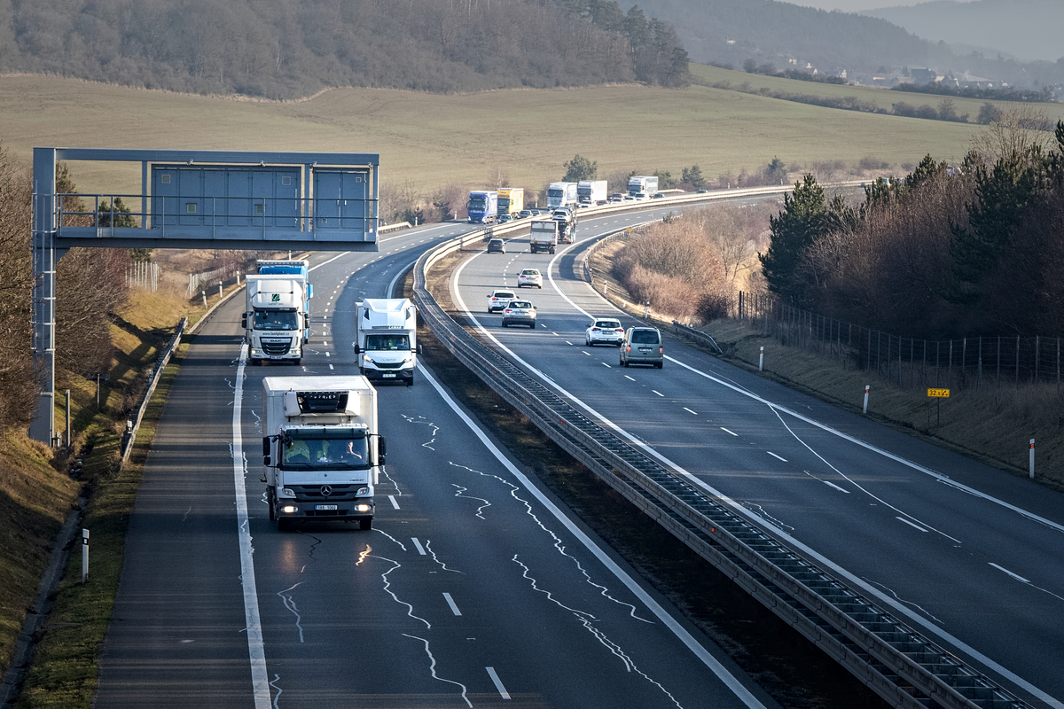 Motorhome among mixed traffic of small HGVs, vans and passenger cars on a Czech motorway curving through rolling hills with toll gantry on the left and km marker 32.5, winter road trip scene, February 2026