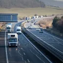 Motorhome among mixed traffic of small HGVs, vans and passenger cars on a Czech motorway curving through rolling hills with toll gantry on the left and km marker 32.5, winter road trip scene, February 2026