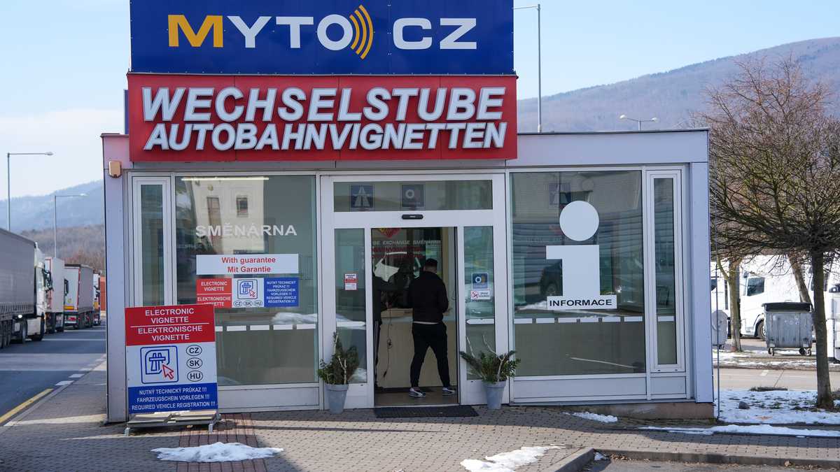 MYTO CZ Wechselstube vignette sales kiosk at a Czech border with German signage, customer at the counter, sells CZ/SK/HU electronic vignettes, HGVs queueing in background, February 2026