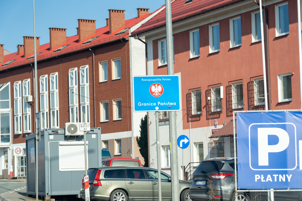 Polish Granica Państwa (State Border) sign with Rzeczpospolita Polska name and Polish white eagle coat of arms at the Kudowa-Zdrój–Náchod border crossing, with former border post buildings, paid parking (płatny) sign and parked cars with DKL Kłodzko district Polish plates, February 2026