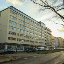 Modern beige SFDI (State Fund of Transport Infrastructure) office building on a Prague Libeň street corner with parked cars, tram tracks, EU and Czech flags by the entrance and "Rekonstrukce Negrelliho viaduktu" branding in the windows, December 2025