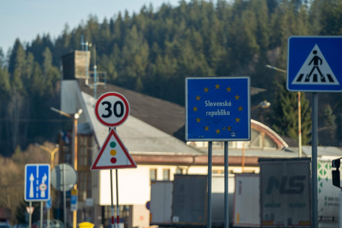 Blue EU-style Slovenská republika border entry sign at the Mosty u Jablunkova–Svrčinovec crossing between Czech Republic and Slovakia, with 30 km/h speed limit, traffic light warning sign and parked HGVs at the former border post in the forested Beskids, February 2026